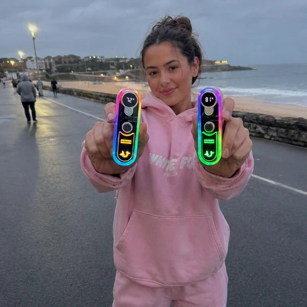 Solis Ember twin hand warmers held up as two separate warmers reading 72° and 81° on their screens, seafront behind