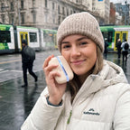 Solis Ember twin hand warmers held against a cheek in cold weather, city trams behind