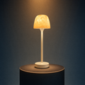 The Solis Vesta table lamp on a lit black podium, warm amber light through its textured dome shade